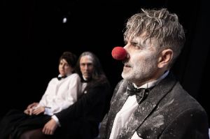 One person wears a red clown’s nose and a suit stained with white dust