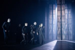 Performers in dark clothing emerge from a dark part of the stage