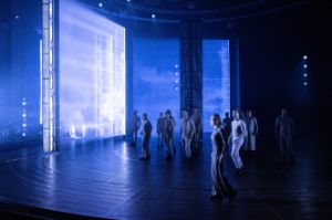 An ensemble of actors stands in a tense posture on a blue-lit stage.