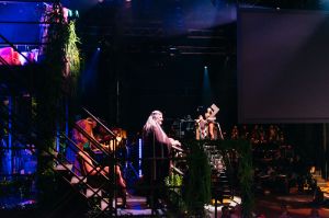 Ein Gerüst auf einer Bühne, darauf Performer*innen und im Hintergrund Publikum