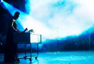 In einem nebligen Raum steht eine Person mit langem Kleid vor einem Rollwagen mit pinken Stöckelschuhen.