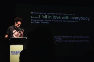 Artist Meloe Gennai has black curly hair and wears sun glasses. He stands at a lectern and reads from a cell phone.  In the background are lines from the text in different font sizes. The following is highlighted in large letters: “I fell in love with everybody.”
