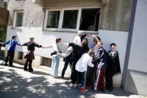 A group of people stand in front of a house wall and try to look into an open window.
