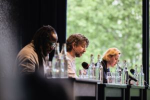 Drei Menschen sitzen nebeneinander an Tischen, vor ihnen Wasserflaschen und Mikrophone.