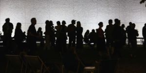 A group of people sit at beer tables on a lawn next to a building in the evening, others queue up at a food truck.