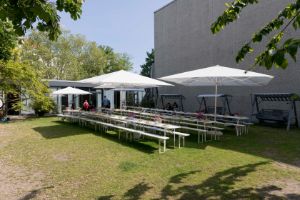 Im Garten des Haus der Berliner Festspiele befinden sich lange weiße Tische mit weißen Bierbänken und weißen Sonnenschirmen.