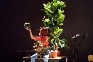 A woman with a flowerpot on her head is surrounded by plants.