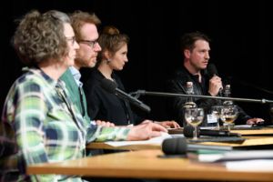 A man speaks seriously into a microphone. Next to him are two women and a man who are also looking seriously towards the audience.