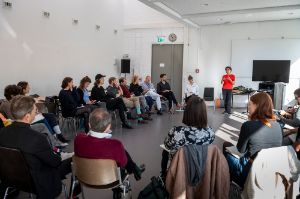 A woman in a red T-shirt speaks in front of a group of attentive listeners.