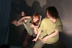 Drei junge Frauen mit dunkel geschminkten Augen in olivfarbenen T-Shirt bewegen sich vor einer weißen Metallwand mit Löchern.