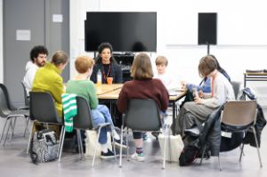 An einem Tisch in einem Konferenzraum sitzen zehn Personen, die ihren Blick auf den Bildschirm im Hintergrund richten, auf dem etwas präsentiert wird.  Daneben ist ein Lautsprecher vor einem Whiteboard erkennbar.