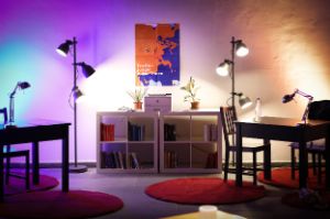 In einem bläulich-gelblich beleuchteten Raum befinden sich zwei Schreibtische auf roten runden Teppichen und zwei quadratische Bücherregale, über denen ein Plakat des Treffen junger Autor*innen zu sehen ist.