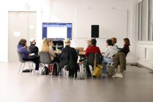 An einem Tisch in einem Konferenzraum sitzen acht Personen, die zum Teil Printmaterialien vor sich liegen haben. Im Hintergrund sind ein großer Bildschirm und ein Lautsprecher erkennbar.