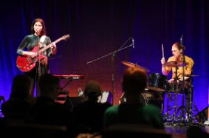 A young woman with a red guitar seems to be singing into a microphone. To her right is a drummer who appears to be playing. Seated spectators are visible in the foreground.