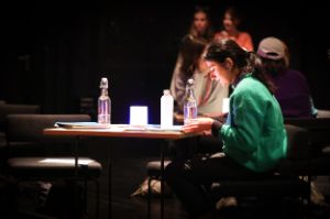 An einem beleuchteten Tisch mit Printmaterialien und Trinkflaschen sitzt eine Teilnehmerin des Treffen junger Autor*innen. Im Hintergrund sitzen und stehen weitere Teilnehmende des Festivals.