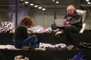 Auf einer stufenartigen Fläche mit Kissen sitzen zwei Teilnehmerinnen des Treffen junger Autor*innen. Eine richtet ihren Blick auf ein Dokument, während die andere an dem Laptop auf ihrem Schoß arbeitet.