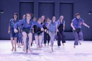 A group of blue-clad young people perform a jumping movement on a stage.