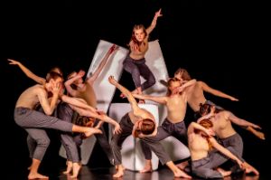 A group of young people wearing grey cloth trousers and beige tops perform movements on a stage. In the middle, a young woman stands on a white platform, pointing and focusing on the viewer.