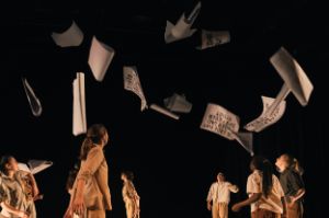 A group of young people, dressed in shades of beige and brown, stand on a stage. Sheets of paper with black lettering fly through the air.