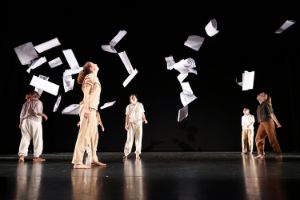 Posters are sailing to the ground against a black background above the heads of the dancers.
