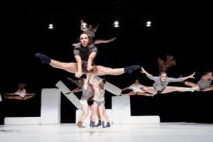 One person hovers above the stage in a straddle jump, behind them are five other dancers in the same pose.