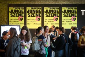 Several people stand in front of a wall with four yellow posters that say “Tanztreffen der Jugend”.