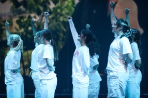 Seven people in white clothes raise their right fists in the air. One person has their mouth taped shut.