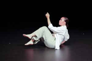 Eine junge Person sitzt in einer Tanzhaltung auf der Bühne, die Beine und einen Arm in die Luft gehoben.