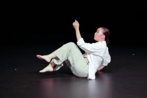 A young person sits on stage in a dancing posture, legs and one arm raised in the air.