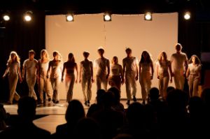 Dreizehn Schauspieler*innen in beiger und weißer Bekleidung vollziehen nebeneinanderstehend, eine Gehbewegung auf einer Bühne.