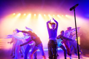 Eine Schauspielerin mit einer schwarzen Lackhose steht mit erhobenen Armen in der Nähe eines Mikrofons an der Spitze einer dreiecksförmigen Formation von weiteren Schauspieler*innen.