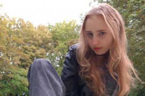 Emily LadyStar hockt vor einem Hintergrund von Bäumen, trägt eine Lederjacke und das lange Haar offen.