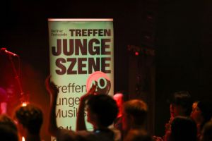 Vor einem grünen Banner mit der Aufschrift „Berliner Festspiele. Treffen junge Szene. Treffen junge Musikszene“ und einem neon-orangenem Button mit einer 40 und der Aufschrift „40 Jahre Treffen junge Musikszene“ stehen einige junge Menschen, die teilweise in die Hände zu klatschen scheinen. Neben dem Banner befindet sich ein Mikrofonständer.