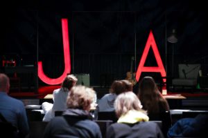 Publikum blickt auf eine Bühne, auf der zwei große Buchstaben „J“ und „A“ rot leuchten.