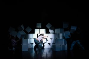 Performer*innen auf der Bühne, eine im Vordergrund durchbricht tanzend eine Mauer aus großen weißen Würfeln.
