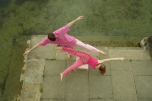 Zwei Performer*innen von oben fotografiert. Sie tragen pinke Kleidung und machen große Bewegungen auf einem Stuhl.