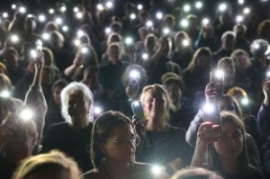 Ein Blick von der Bühne ins Publikum. Viele Personen im Publikum gucken neugierig und halten ihre Handys mit eingeschalteter Taschenlampe hoch.