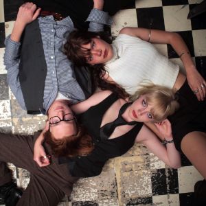 The three band members lie on a floor with a chequerboard pattern and look into the camera.