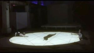 A circle of light on a stage in which two people stretch out their hands to the left and right