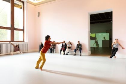 In einem hellrosa gestrichenen Raum performen mehrere Menschen. Das Publikum sitzt auf Bänken und beobachtet die Bewegungen der tanzenden Menschen.
