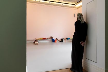 In einem hellrosa gestrichenen Raum findet eine Live-Performance mit dem Titel „study now steady“ statt. Mehrere Tänzer:innen bewegen sich dynamisch durch den Raum und beziehen dabei den Boden, die Wände und die Bänke in ihre Choreografie ein. Eine Person steht in einem Türrahmen und beobachtet aufmerksam das Geschehen.