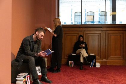 In der Ecke eines Raumes mit Teppichboden aus dunkelrotem Samt, befindet sich ein Bereich mit Büchern. In dieser Bücherecke stöbern Besucherinnen der Ausstellung „Ligia Lewis: I’M NOT HERE FORRRRR…“ in zur Ausstellung thematisch passender Literatur.