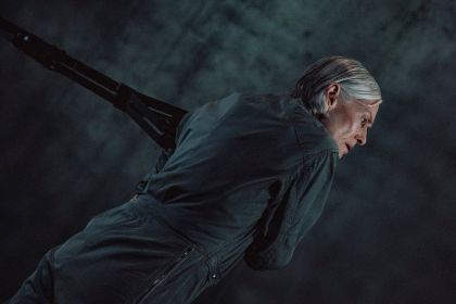Lucinda Childs pulls on a rope. The choreographer wears a dark grey jumpsuit and has grey hair.