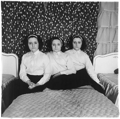 Drei weiblich gelesene Personen sitzen auf einem Bett. Die Personen sind Drillinge, tragen die gleiche Kleidung und die gleiche Frisur.