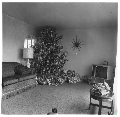 In der Ecke eines schlicht möblierten Wohnzimmers steht ein deckenhoher, brachtvoll geschmückter Weihnachtsbaum. Unter ihm unzählige Geschenke.