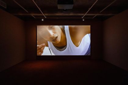 A screen hangs in an empty room, showing a close-up of the upper body of a female-presenting person.