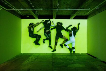 A screen hangs in an empty room, showing four people lying in unusual poses, surrounded by bright green light.