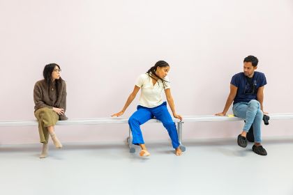 Three people sit evenly spaced on a narrow white bench, set against a pink background.