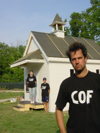Christoph Schlingensief steht vor einer weißen, aus Holz gefertigten Kapelle. Hinter ihm stehen zwei Personen, sie alle tragen T-Shirts mit dem Schriftzug „COF“.