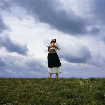 Eine Person mit einem schwarzen Tuch auf dem Kopf massiert sich im Freien ihre nackten Brüste.